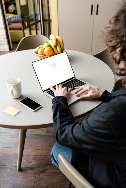 KYIV, UKRAINE - APRIL 25, 2020: back view of freelancer using laptop with Google on screen near smartphone, coffee cup, sticky notes and fruits - Photo, image