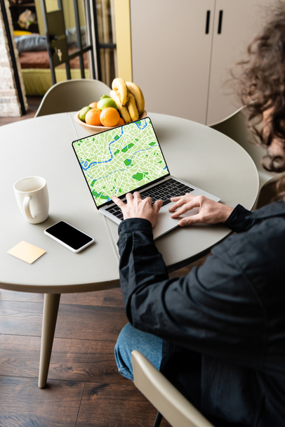back view of freelancer using laptop with map on screen near smartphone, coffee cup, sticky notes and fruits - Foto, afbeelding
