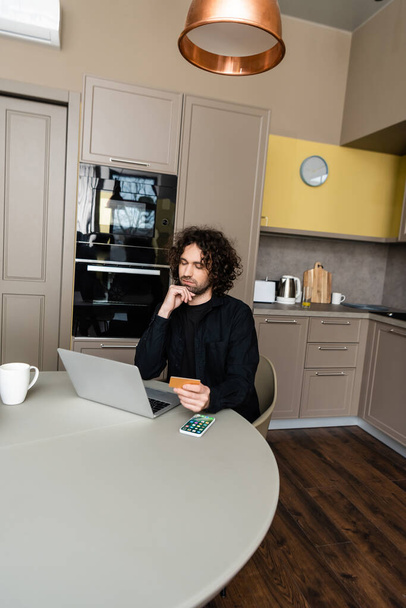 KYIV, UKRAINE - APRIL 25, 2020: thoughtful freelancer holding credit card while looking at laptop near Apple smarphone with homepage on screen - Valokuva, kuva