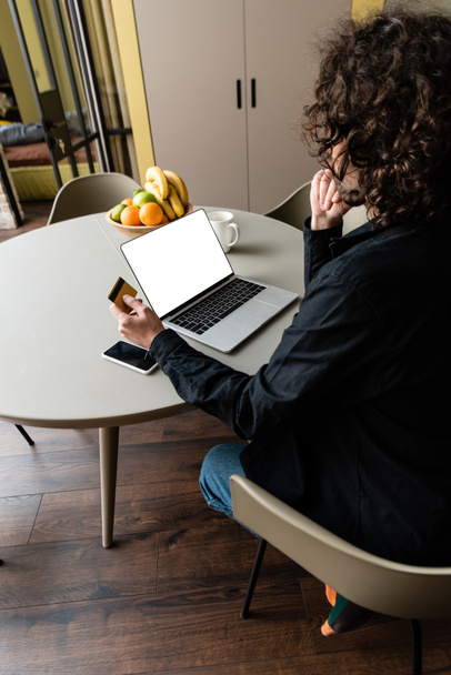 vista posterior del freelancer que sostiene la tarjeta de crédito mientras que está sentado en el ordenador portátil con la pantalla blanca, smartphone y frutas
 - Foto, Imagen