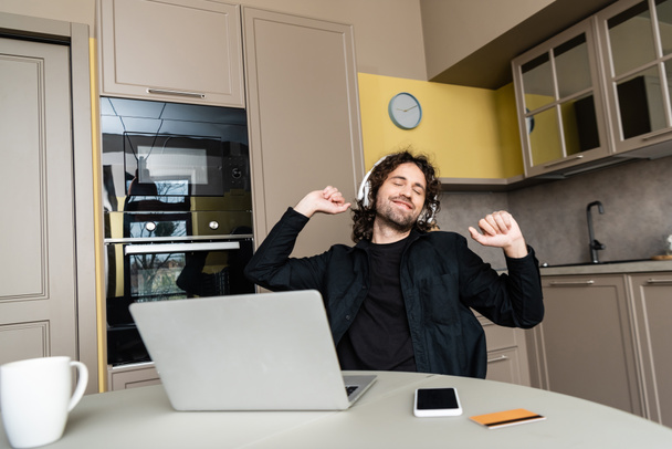 Επιλεκτική εστίαση του χαμογελαστού ανθρώπου στα ακουστικά ακούγοντας μουσική κοντά σε gadgets και πιστωτική κάρτα στο τραπέζι της κουζίνας  - Φωτογραφία, εικόνα