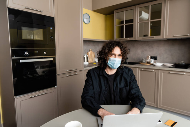 Hombre en máscara médica utilizando el ordenador portátil cerca de teléfono inteligente y tarjeta de crédito en la mesa de la cocina
  - Foto, Imagen