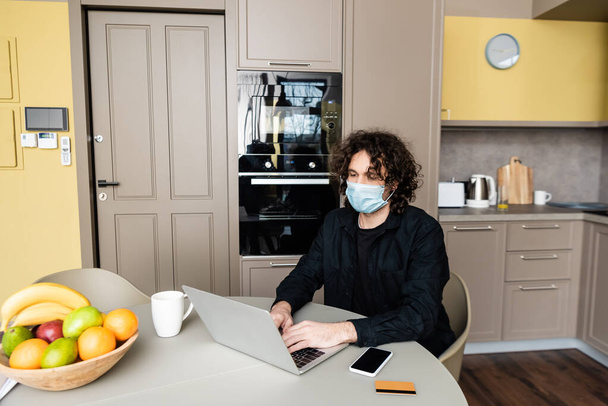 Man in medical mask using laptop near smartphone, credit card and fruits on kitchen table  - Photo, image