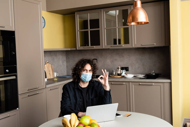 Hombre en máscara médica mostrando gesto ok cerca de gadgets y tarjeta de crédito en la mesa de la cocina
  - Foto, imagen