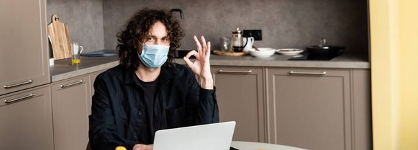 Panoramic shot of man in medical mask showing okay gesture near laptop in kitchen  - 写真・画像