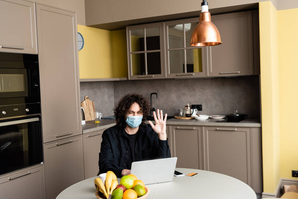 Man in medical mask having video call on laptop near smartphone and credit card on table in kitchen  - Valokuva, kuva