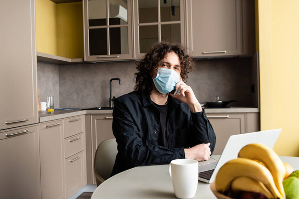 Selective focus of freelancer in medical mask talking on smartphone near laptop, fruits and cup on kitchen table  - 写真・画像