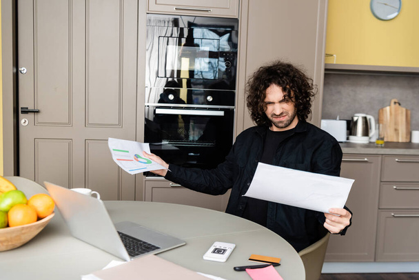 KYIV, UKRAINE - APRIL 25, 2020: Selective focus of confused freelancer holding documents near laptop and smartphone with uber app in kitchen  - Foto, afbeelding