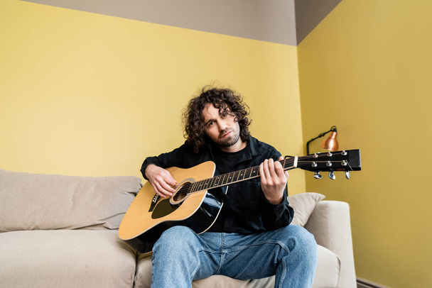 Hombre guapo mirando a la cámara mientras sostiene la guitarra acústica en el sofá
  - Foto, Imagen