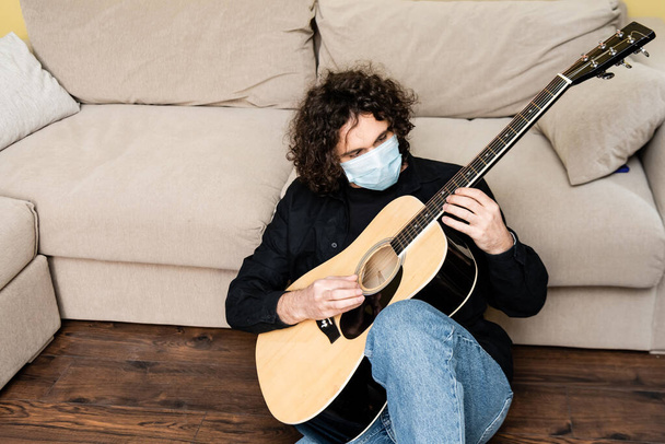 Homem de máscara médica tocando guitarra acústica no chão em casa
  - Foto, Imagem