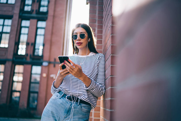 Millennial hipster girl in sunglasses standing outdoors and writing text review publication about popular music set for sharing to social networks, young female listening audio book and messaging - Foto, afbeelding