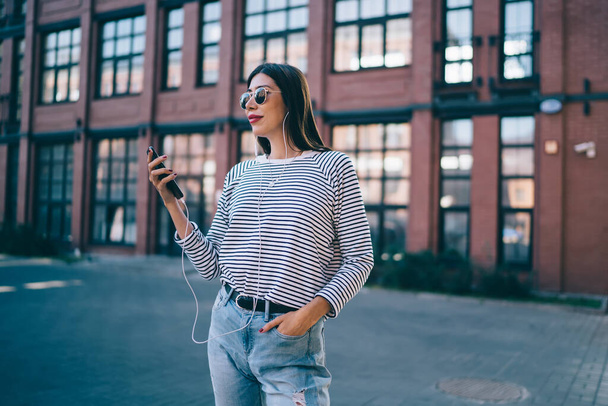 Carefree female teenager in trendy sunglasses using electronic headphones for listening radio music during leisure time in city, Latino millennial woman enjoying media set with popular audio tracks - Foto, imagen