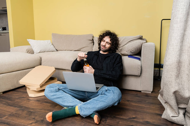 Freelancer opent fles bier in de buurt van laptop en pizza dozen op de vloer  - Foto, afbeelding