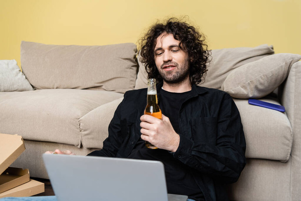 Selective focus of freelancer holding bottle of beer and using laptop near pizza boxes and couch at home  - Foto, Imagen