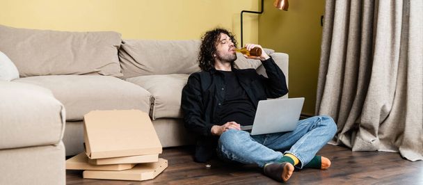 Colpo panoramico di freelance bere birra vicino laptop e scatole di pizza sul pavimento
  - Foto, immagini