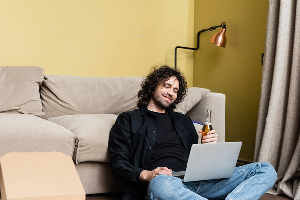 Uśmiechnięty wolny strzelec trzymający butelkę piwa w pobliżu laptopa i pudełka po pizzy w domu  - Zdjęcie, obraz