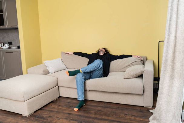 Curly man looking up while sitting on couch in living room - Photo, Image