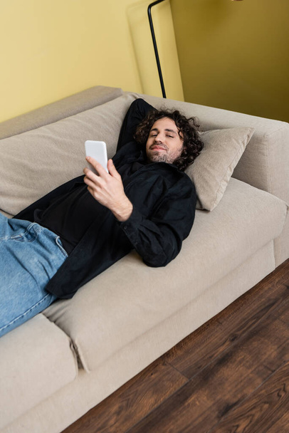 Curly man looking at camera and holding smartphone on couch  - Zdjęcie, obraz