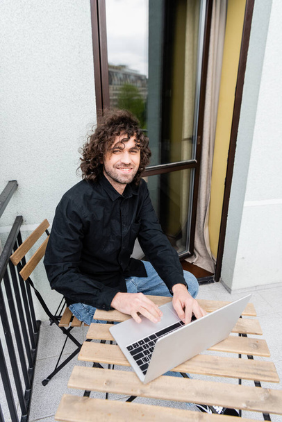 Красивый мужчина улыбается в камеру во время использования ноутбука на балконе дома
  - Фото, изображение