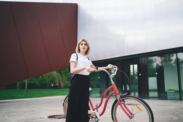 Attractive dreamy young blonde woman in casual outfit with small backpack standing on street holding handlebar of vintage bicycle looking away  - Foto, Imagem