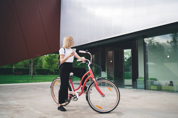Вид сбоку на симпатичную женщину в повседневной одежде, внимательно пользующуюся смартфоном, сидя на велосипеде на городской улице и отводя взгляд - Фото, изображение
