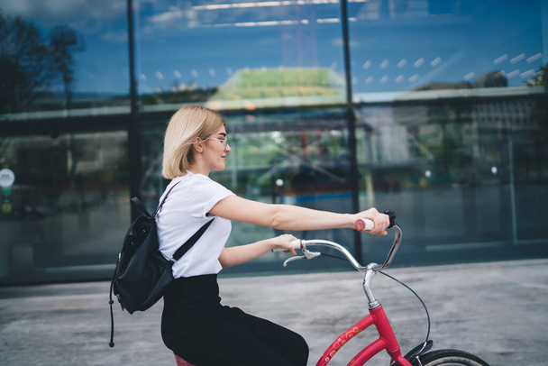 Side view of young pretty blonde woman in casual clothes with black backpack sitting on bicycle and riding on street - Φωτογραφία, εικόνα