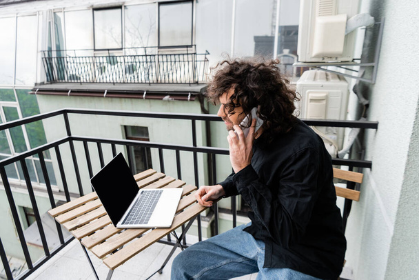 Teleworker talking on smartphone near laptop on table on balcony  - Foto, imagen