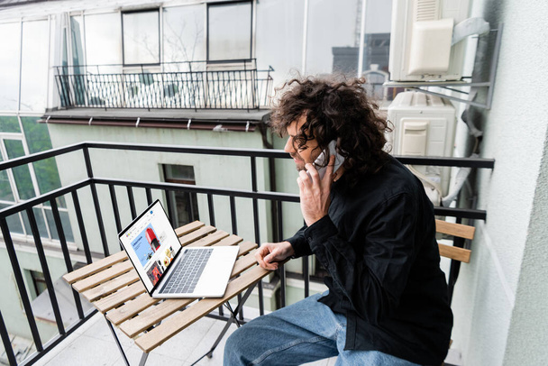 KYIV, UKRAINE - APRIL 25, 2020: Freelancer talking on smartphone near laptop with ebay website on balcony  - Zdjęcie, obraz