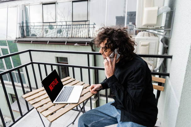 KYIV, UKRAINE - APRIL 25, 2020: Curly man talking on smartphone near laptop with netflix website on balcony  - Photo, image