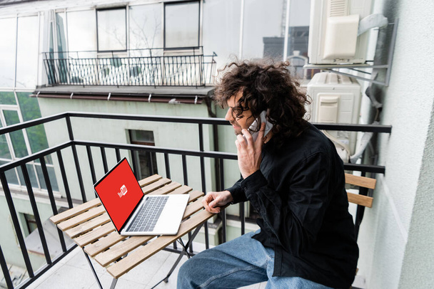 KYIV, UKRAINE - APRIL 25, 2020: Freelancer talking on smartphone near laptop with youtube website on table on balcony  - Valokuva, kuva
