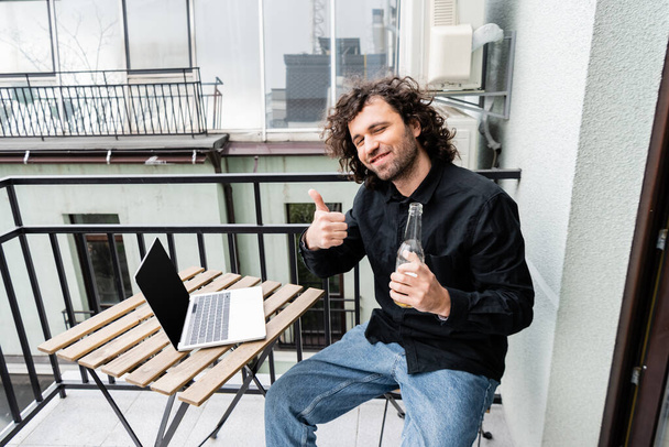 Sonriente hombre mostrando como gesto mientras sostiene la botella de cerveza cerca de la computadora portátil en el balcón
  - Foto, imagen