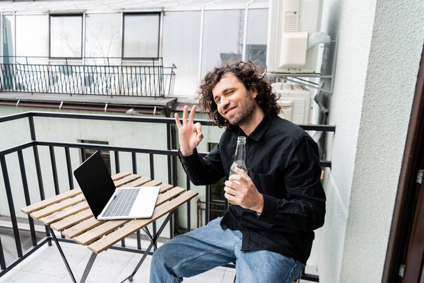 Freiberufler lächelt in die Kamera und zeigt eine freundliche Geste, während er auf dem Balkon eine Bierflasche neben Laptop hält  - Foto, Bild