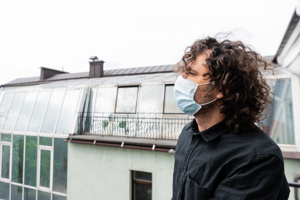 Πλευρική άποψη του σγουρά άνθρωπος στην ιατρική μάσκα κοιτάζοντας μακριά, ενώ στέκεται στο μπαλκόνι  - Φωτογραφία, εικόνα