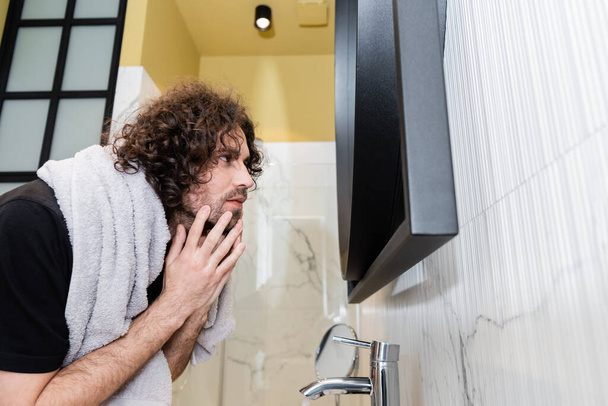 Вид збоку красивий кучерявий чоловік торкається обличчя, дивлячись на дзеркало у ванній
 - Фото, зображення
