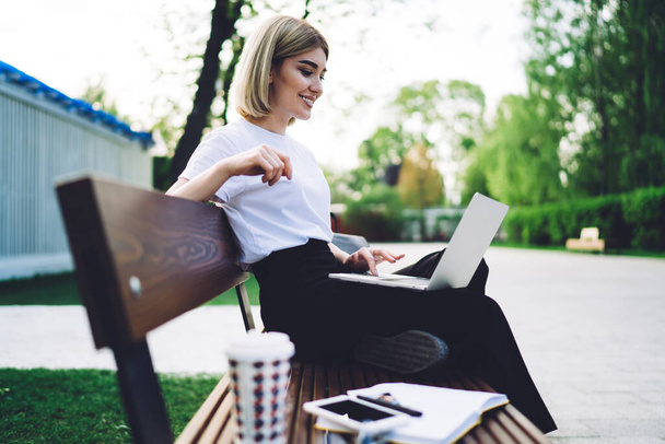 Вид сбоку на жизнерадостную и оптимистичную женщину с улыбкой, сидящую на скамейке в парке и чиллирующую за чашкой кофе и просмотром социальных сетей  - Фото, изображение