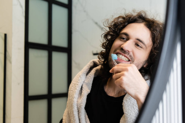 Selective focus of curly man with towel on shoulders brushing teeth in bathroom - Фото, изображение