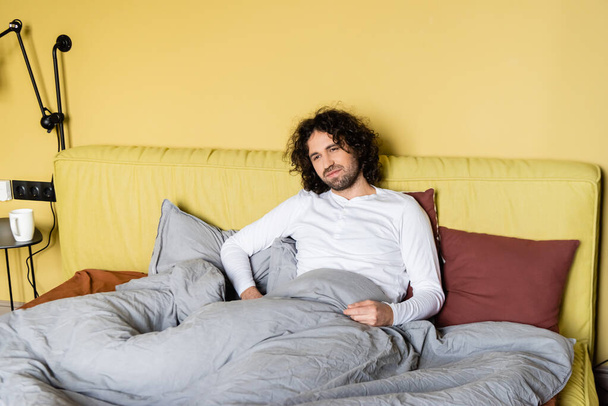 Komea kihara mies istuu sängyllä kotona
  - Valokuva, kuva