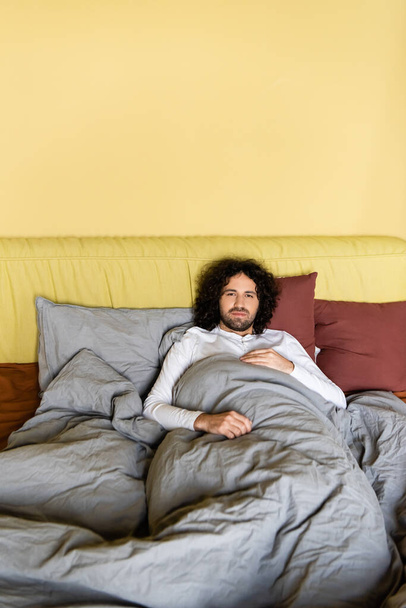 Curly man looking at camera while lying on bed  - Foto, Imagen