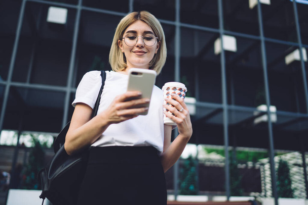 From below smiling adult elegant female with handbag and in glasses focusing and interacting with phone while standing and holding takeaway hot drink on street near office building - 写真・画像