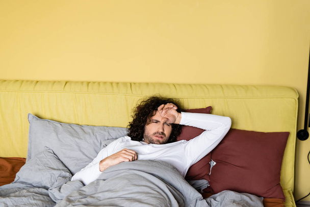 Αναστατωμένος άντρας με το χέρι κοντά στο μέτωπο ξαπλωμένος στο κρεβάτι στο σπίτι  - Φωτογραφία, εικόνα