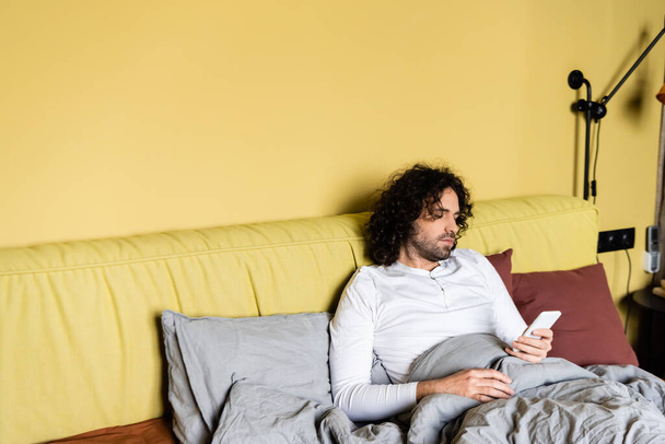 Handsome curly man using smartphone on bed  - Fotó, kép