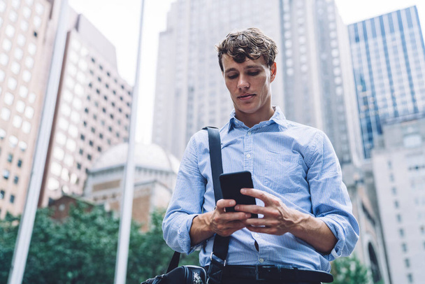 Young businessman in shirt with leather bag messaging on phone walking on the backend of high buildings of New York - 写真・画像