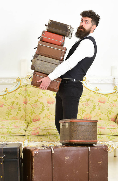 Macho, elegant porter on strict face carries pile of vintage suitcases. Man with beard and mustache wearing classic suit delivers luggage, luxury white interior background. Butler and service concept. - Photo, image