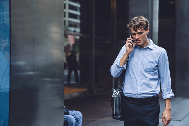 Zelfverzekerde jongeman in formele outfit met leren tas praten op mobiele tijdens het lopen op straat van New York City  - Foto, afbeelding