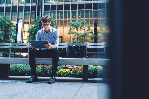 Handsome thoughtful young male in glasses sitting on bench in front of office building using tablet on street in Manhattan, New York city - 写真・画像