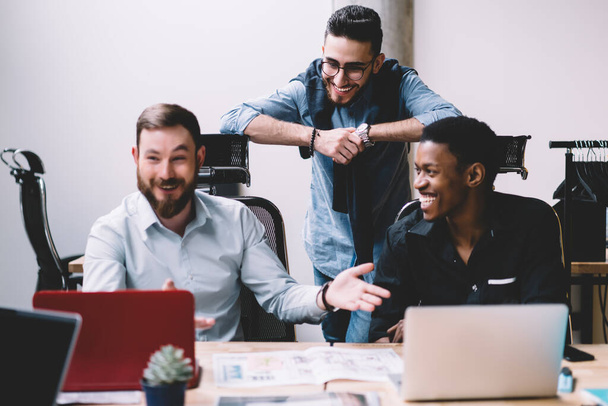 Diverse male businesspeople dressed in shirts laughing while having lively conversation about startup project using laptops sitting in bright office - Foto, imagen