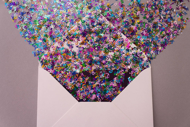 Opened white envelope with multicolored sparkle glitter confetti letters on pastel lilac background. Festive greeting concept. Flat lay style. Top view - 写真・画像