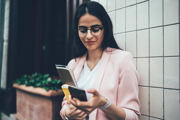 Modern busy woman in eyeglasses and blazer reading urgent email from client on smartphone while standing next to wall of city building with papers and diary in hand  - Foto, Bild