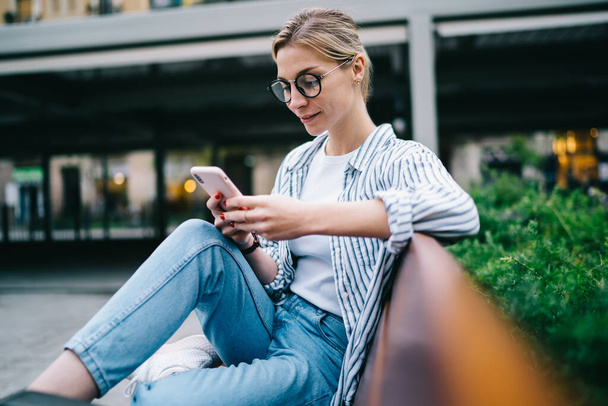Side view of attractive blond woman in casual clothes with round eyeglasses using modern smartphone and smiling while sitting on bench near green bush on blurred street background - Photo, image