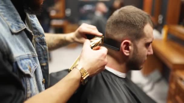 Trendy barber cuts bearded mans hair with a clipper in barbershop. Mens hairstyling and hair cutting in salon. Grooming the hair with trimmer. Hairdresser doing haircut in retro hair salon. - Felvétel, videó